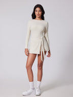 Woman wearing a white long-sleeve dress with a tie detail on a plain background
